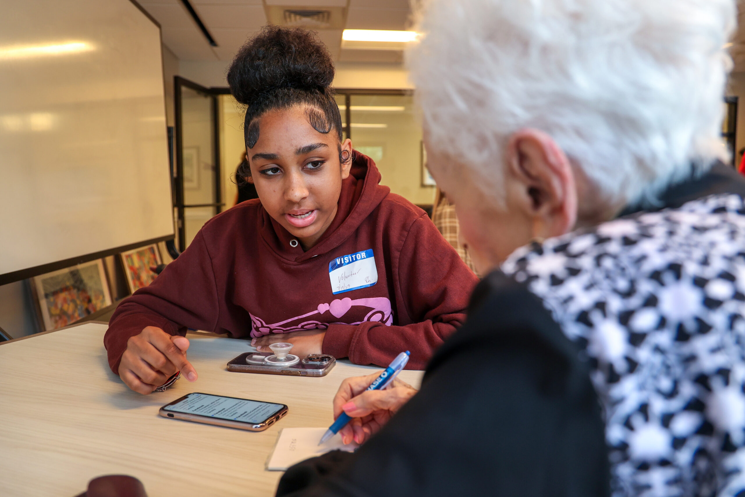 A teen points at a phone on a table while speaking with a senior woman