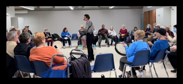 A woman stands in the middle of a group of people sitting in chairs, participating in a drum circle