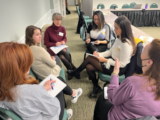 A group of six younger women sit in a meeting hall writing on a paper