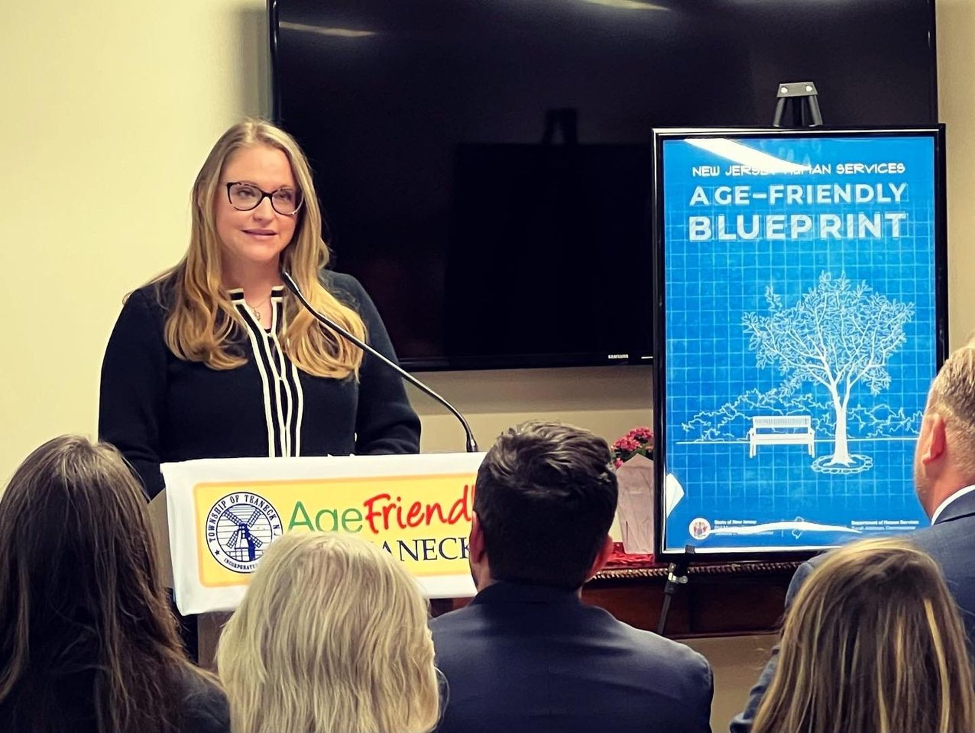 A blonde woman presenting an Age-Friendly Blueprint to a crowd at an Age-Friendly Teaneck meeting